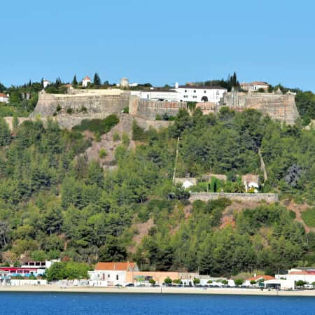 Fortress on a hillside with trees and buildings below, overlooking water.