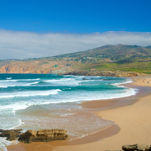 Beach scene with waves, sand, and mountains under a blue sky.