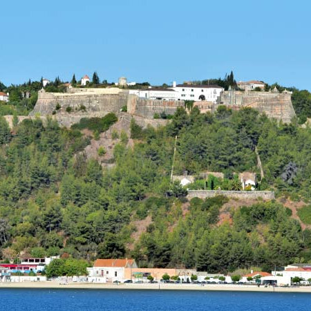 Fortress on a hillside with trees and buildings below, overlooking water.